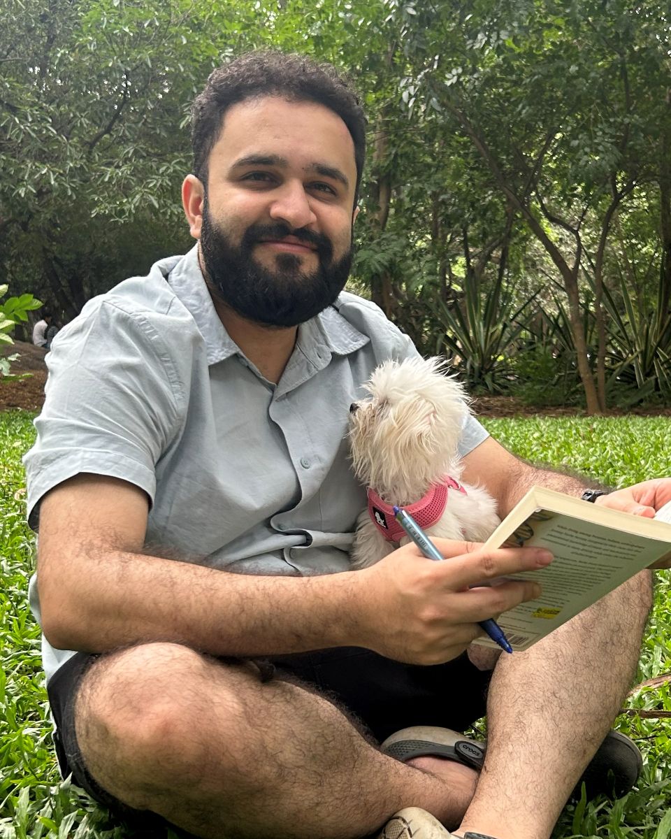 Akshat Kapur sitting on the grass in a park, holding a book, with a small white dog